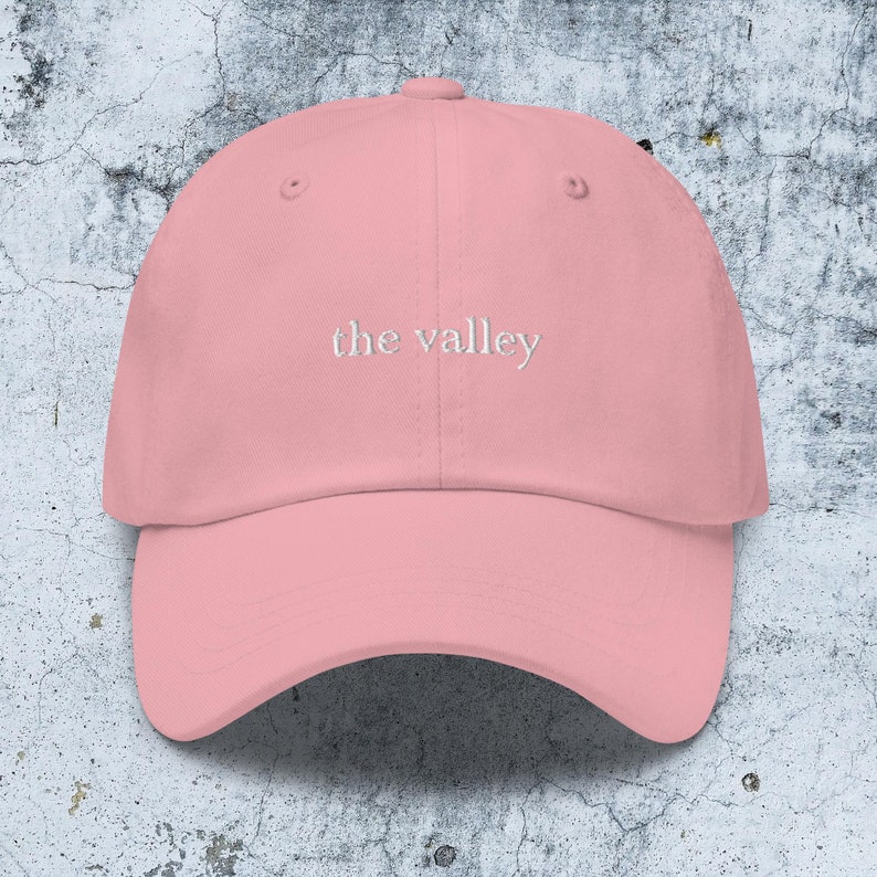 May include: A light pink baseball cap featuring the text "the valley" embroidered in white thread across the front center. The cap has a structured crown, a curved brim, and a button at the top, showcasing a clean and simple design.