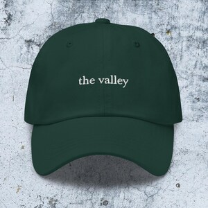May include: A dark forest green baseball cap featuring the text the valley embroidered in white thread across the front center. The cap has a structured crown, a curved brim, and a button at the top, showcasing a clean and simple design.
