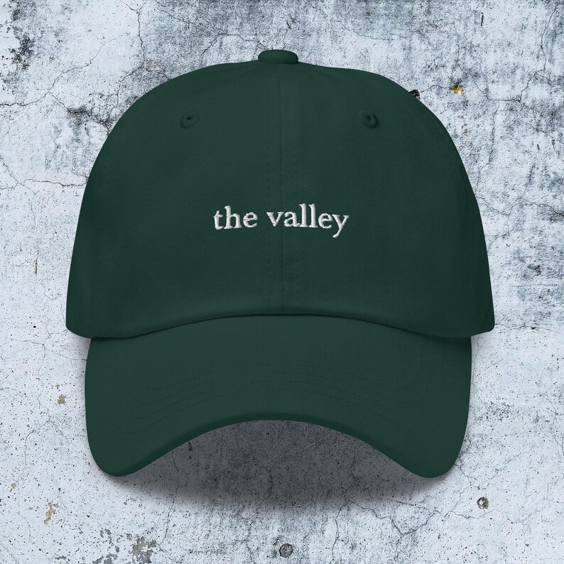 May include: A dark forest green baseball cap featuring the text the valley embroidered in white thread across the front center. The cap has a structured crown, a curved brim, and a button at the top, showcasing a clean and simple design.