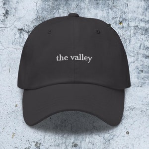 May include: A charcoal gray baseball cap featuring the text the valley embroidered in white thread across the front center. The cap has a structured crown, a curved brim, and a button at the top, showcasing a clean and simple design.