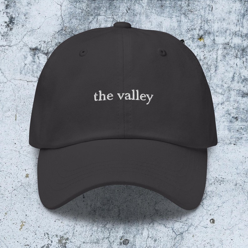 May include: A charcoal gray baseball cap featuring the text the valley embroidered in white thread across the front center. The cap has a structured crown, a curved brim, and a button at the top, showcasing a clean and simple design.