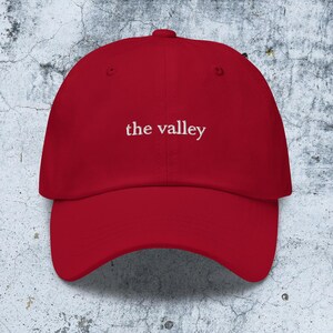 May include: A red baseball cap featuring the text "the valley" embroidered in white thread across the front center. The cap has a structured crown, a curved brim, and a button at the top, showcasing a classic six-panel design with ventilation eyelets.