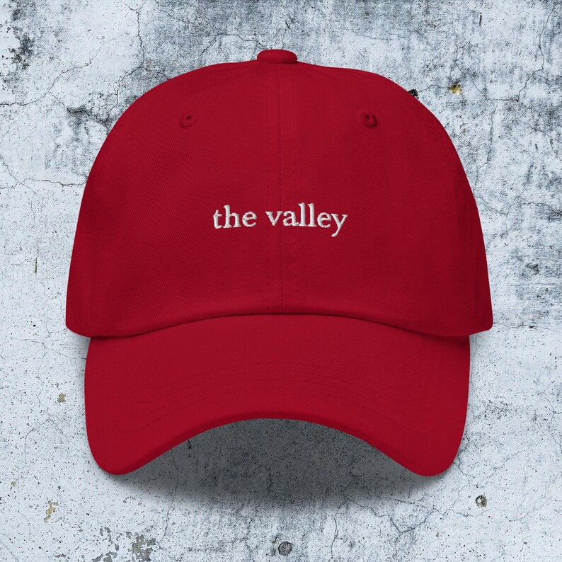 May include: A red baseball cap featuring the text "the valley" embroidered in white thread across the front center. The cap has a structured crown, a curved brim, and a button at the top, showcasing a classic six-panel design with ventilation eyelets.