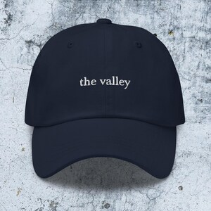 May include: A navy blue baseball cap featuring the text the valley embroidered in white thread across the front center. The cap has a structured crown, a curved brim, and a button at the top, presenting a clean and simple design for headwear.