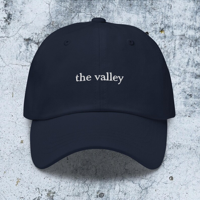 May include: A navy blue baseball cap featuring the text the valley embroidered in white thread across the front center. The cap has a structured crown, a curved brim, and a button at the top, presenting a clean and simple design for headwear.