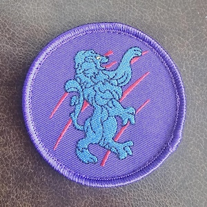 Op de afbeelding: Paarse geborduurde patch met een blauw leeuwenontwerp. De leeuw staat op zijn achterpoten met zijn klauwen uitgespreid. De achtergrond is paars met rode stiksels.