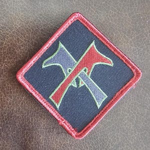 May include: A black patch with red and green embroidered crossed axes. The patch has a red border.