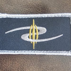 Puede incluir: Un parche bordado gris con un logotipo estilizado blanco y dorado. El logotipo presenta una letra "S" estilizada con un diseño geométrico dorado en el centro.