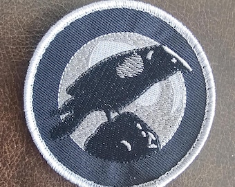 Puede incluir: Un parche bordado en blanco y negro que representa un cuervo posado en una roca, con una luna en el fondo. El parche es circular y tiene un borde blanco.