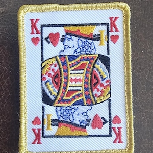 Könnte beinhalten: Ein weißer gestickter Aufnäher mit goldener Umrandung, der die Spielkarte König der Herzen zeigt. Die Karte hat in jeder Ecke ein rotes Herz-Symbol und den Buchstaben 'K' in der oberen rechten und unteren linken Ecke.