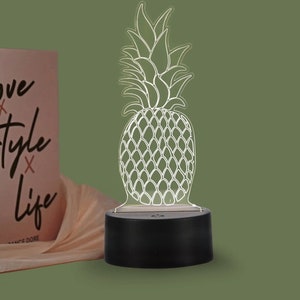 Puede incluir: Una luz nocturna con forma de piña blanca con una base negra. La piña está delineada en blanco y tiene un diseño detallado.