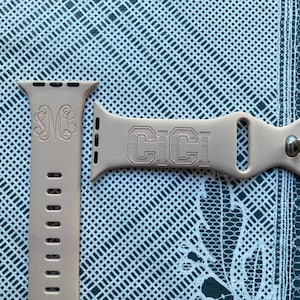 May include: Two light pink Apple Watch bands. One band features the engraved letters "MB" in a decorative font. The other band has the engraved letters "CICI". Both bands are made of a flexible material and have a metal clasp.