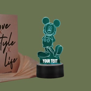 May include: A teal-colored Mickey Mouse-shaped LED night light. The light has a black base with the words "YOUR TEXT" displayed. A book with the words "Love Style Life" is in the background.
