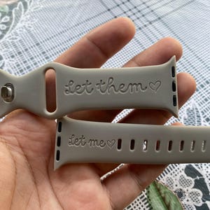 May include: Two light gray watch bands with the phrases "Let them" and "Let me" engraved, each with a heart symbol. The bands are made of a flexible material and have a series of holes for adjustment.