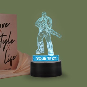 Pode incluir: Uma luz noturna impressa em 3D azul que apresenta um soldado futurista segurando um rifle. A base da luz é preta e tem uma pequena placa retangular com o texto "YOUR TEXT" nela.