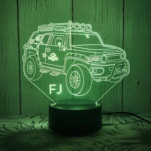May include: A green LED night light shaped like a Toyota FJ Cruiser with a roof rack and the letters "FJ" below the car. The night light is sitting on a black base.