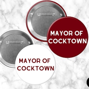 May include: Two circular pin-back buttons featuring the text MAYOR OF COCKTOWN. One button has a deep red background with white text, and the other has a white background with deep red text. Both buttons have a silver metal back with a pin.