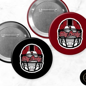 May include: Four round pinback buttons. Two buttons are black, and two are burgundy. Each button features a football helmet graphic with the text "Stay Cocky" and "Since 1895". The buttons have a silver metal backing.