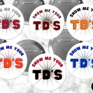 May include: A collection of white round buttons with the text "SHOW ME YOUR TD'S" in various colors, including red, orange, and blue. The buttons have a radial design emanating from the center.