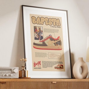 May include: A framed poster with a vintage design featuring a brown and white sneaker with a red and yellow star on the side. The poster has the text "BAPESTA x Kanye West" on it. The poster is hanging on a wall above a wooden cabinet with a stereo on top.