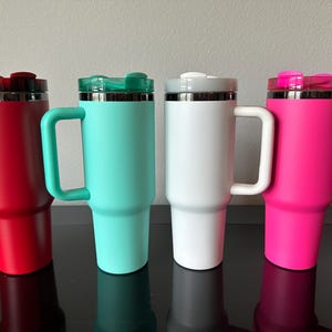 May include: Four insulated travel mugs in red, teal, white, and pink, each with a handle and a lid. The mugs are cylindrical with a tapered base and a stainless steel rim. The handles and lids match the body colour.