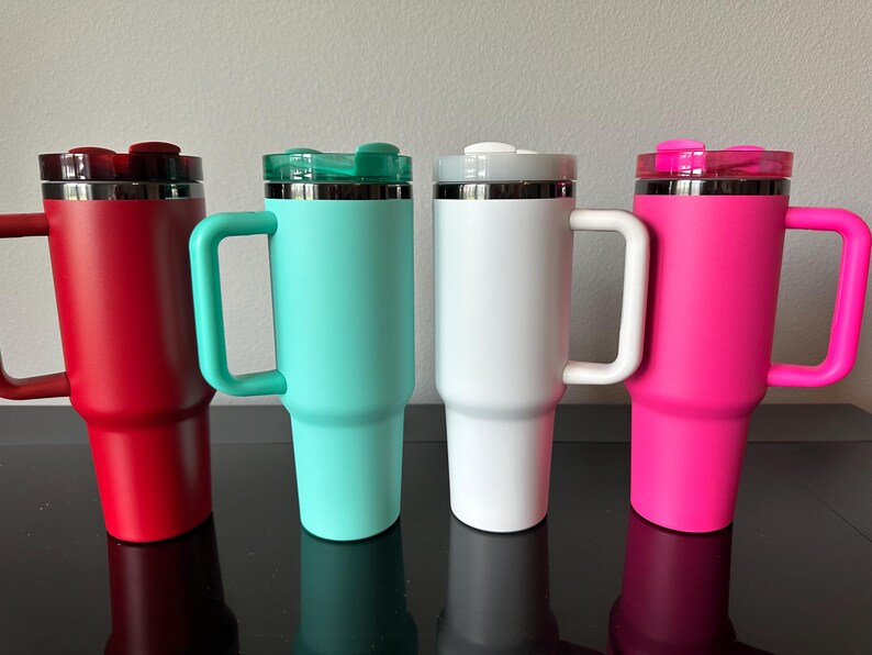 May include: Four insulated travel mugs in red, teal, white, and pink, each with a handle and a lid. The mugs are cylindrical with a tapered base and a stainless steel rim. The handles and lids match the body colour.