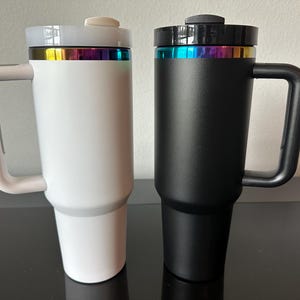May include: Two insulated travel mugs, one white and one black, with a handle and a lid. Each mug has a rainbow-coloured band near the top. The mugs are designed to keep beverages hot or cold for extended periods.