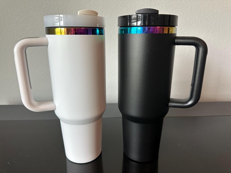 May include: Two insulated travel mugs, one white and one black, with a handle and a lid. Each mug has a rainbow-coloured band near the top. The mugs are designed to keep beverages hot or cold for extended periods.