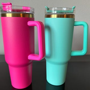 May include: Two insulated tumblers with handles. One is bright pink, the other is teal. Both have a gold band near the top and a clear plastic lid with a flip-top closure. The handles and flip-top closures match the tumbler's colour.