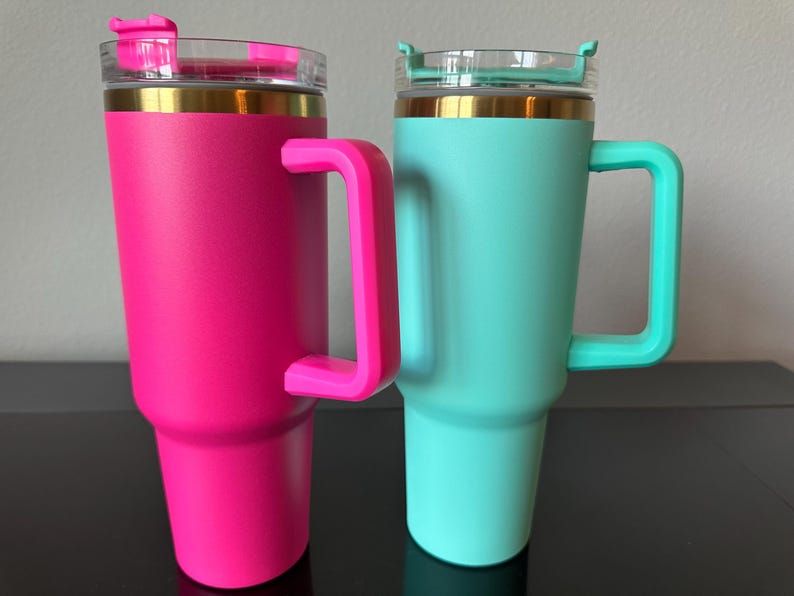 May include: Two insulated tumblers with handles. One is bright pink, the other is teal. Both have a gold band near the top and a clear plastic lid with a flip-top closure. The handles and flip-top closures match the tumbler's colour.