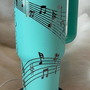 May include: A teal insulated tumbler with a handle, featuring a musical staff design with notes and the name "Deanna" in a cursive font. The tumbler is on a black base.