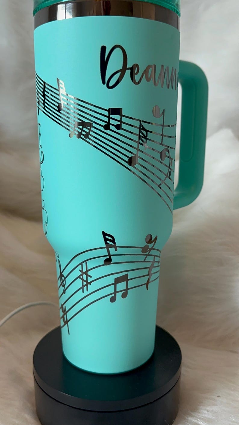 May include: A teal insulated tumbler with a handle, featuring a musical staff design with notes and the name "Deanna" in a cursive font. The tumbler is on a black base.