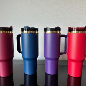 May include: Four insulated travel mugs in shades of red, blue, and purple. Each mug has a handle, a black lid with a gold rim, and a matching coloured body. The mugs are designed to keep beverages hot or cold.