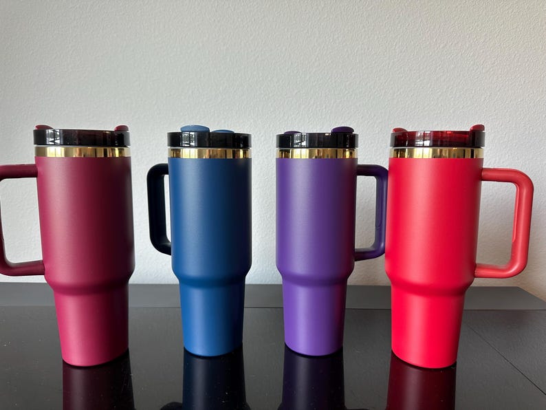 May include: Four insulated travel mugs in shades of red, blue, and purple. Each mug has a handle, a black lid with a gold rim, and a matching coloured body. The mugs are designed to keep beverages hot or cold.