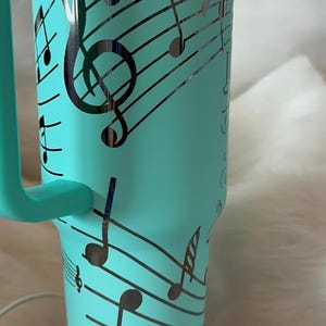 May include: A teal insulated tumbler with a handle, decorated with black musical notes and staff lines. The design wraps around the tumbler, featuring treble clefs and various note symbols. The tumbler sits on a black base.