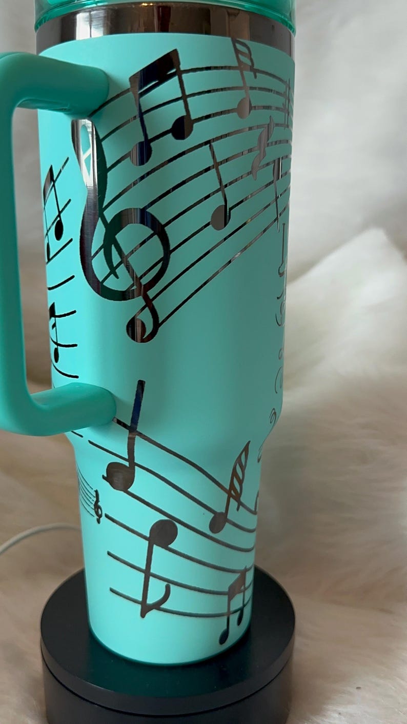 May include: A teal insulated tumbler with a handle, decorated with black musical notes and staff lines. The design wraps around the tumbler, featuring treble clefs and various note symbols. The tumbler sits on a black base.