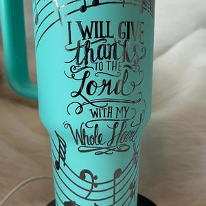 May include: A teal travel mug with a handle, decorated with musical notes and the words "I WILL GIVE thanks TO THE Lord WITH MY Whole Heart". The mug is on a black base.