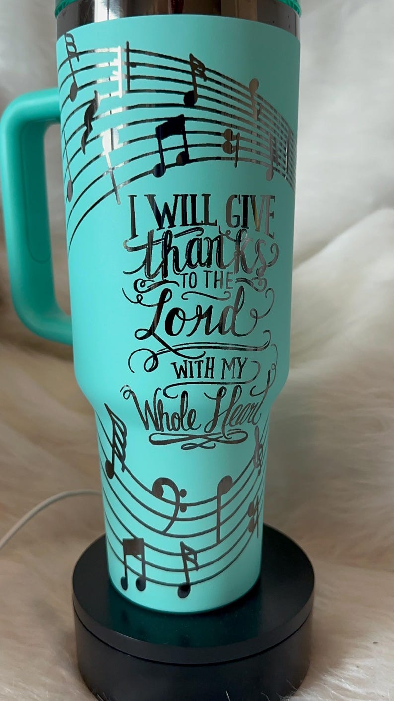 May include: A teal travel mug with a handle, decorated with musical notes and the words "I WILL GIVE thanks TO THE Lord WITH MY Whole Heart". The mug is on a black base.