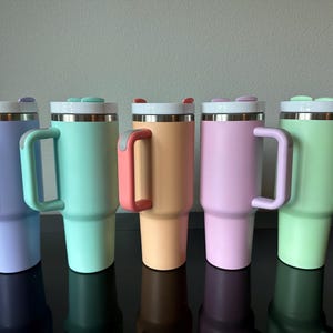 May include: Five pastel-coloured insulated tumblers: lavender, mint, peach, pink, and light green. Each has a stainless steel rim, a white lid with a small button, and a matching handle. Designed to keep drinks cold or hot.