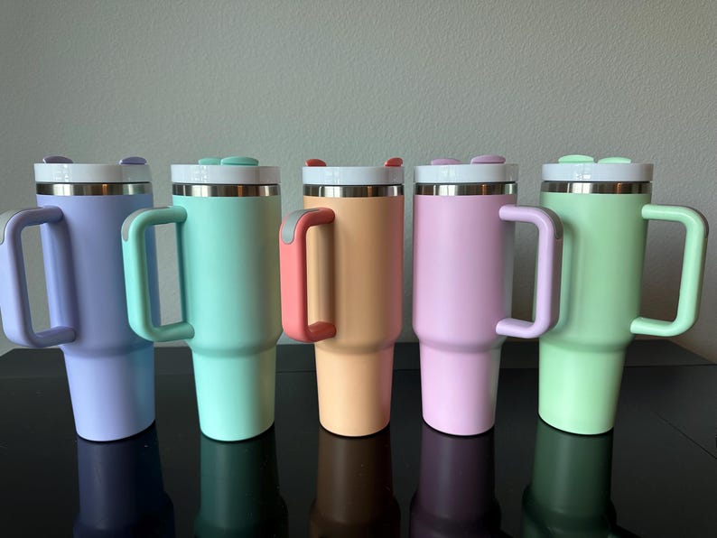 May include: Five pastel-coloured insulated tumblers: lavender, mint, peach, pink, and light green. Each has a stainless steel rim, a white lid with a small button, and a matching handle. Designed to keep drinks cold or hot.