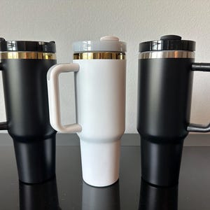 May include: Three insulated travel mugs with handles. Two are black with gold trim, and one is white with gold trim. Each mug has a black or grey lid. The mugs are designed to keep beverages hot or cold.
