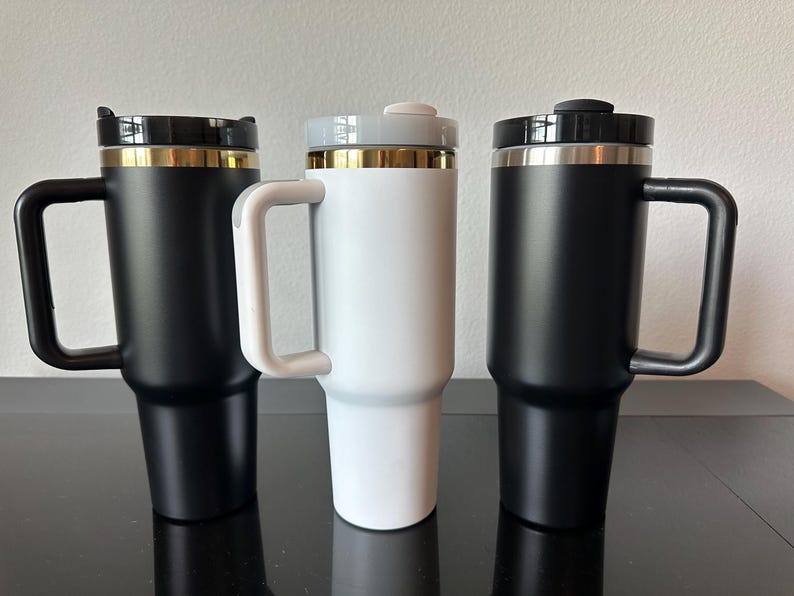 May include: Three insulated travel mugs with handles. Two are black with gold trim, and one is white with gold trim. Each mug has a black or grey lid. The mugs are designed to keep beverages hot or cold.