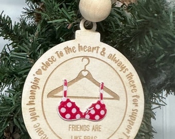 May include: Wooden Christmas ornament with a red and white polka dot bra design. The ornament has the text "Friends are like bras" and other text around the edge. It hangs from a string with two wooden beads.