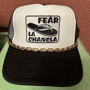 May include: A black and white trucker hat with a gold chain trim. The hat has a white front panel with a black and white patch that says "FEAR LA CHANCLA" and a black flip-flop graphic.