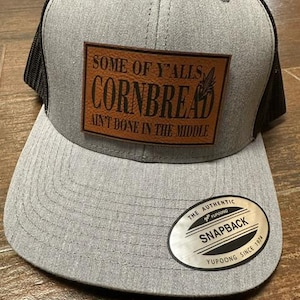 May include: Gray and black trucker hat with a brown leather patch that says "Some of y'alls cornbread ain't done in the middle". The hat has a black mesh back and a silver YUPOONG SNAPBACK label.