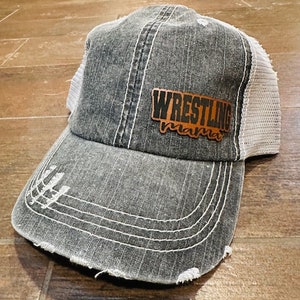 May include: Distressed gray trucker hat with a white mesh back and a curved brim. The hat features a brown leather patch with the words "Wrestling Mama" in a stylized font. White stitching accents the hat's design.