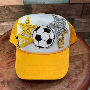 May include: A yellow and white trucker hat with a soccer ball, two stars, and a 'Game Day' pennant patch.