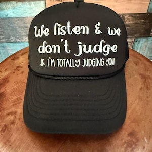 May include: Black trucker hat with white text that reads "We listen & we don't judge. JK. I'm totally judging you!"