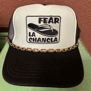 May include: A black and white trucker hat with a gold chain trim. The hat has a white front panel with a black and white patch that says "FEAR LA CHANCLA" and a black flip-flop graphic.