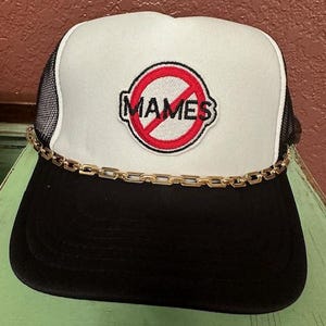 May include: A black and white trucker hat with a red circle and the word "MAMES" in white letters. The hat has a gold chain around the brim.
